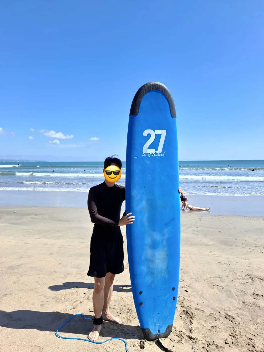 ‘27 Surf School’이라 적힌 파란 서프보드를 세워 잡고 해변에 선 모습
